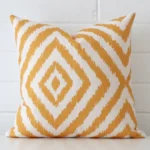 Gold patterned cushion cover in front of a white wall. It has a square size and is made from a linen material.